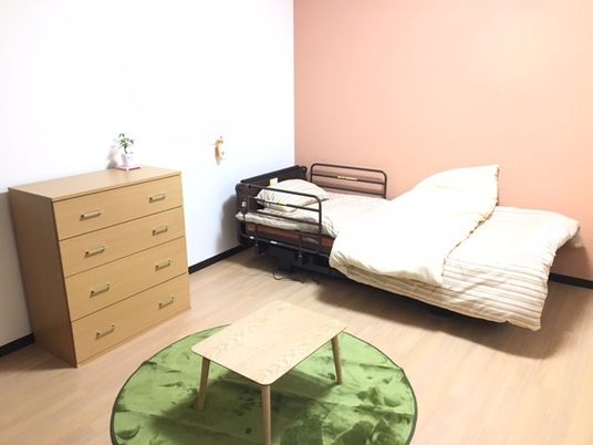 居室の壁は白と暖色系の２色使いで明るい雰囲気。部屋の中にはベッドとタンス、フローリングに敷かれたラグの上には小さなテーブルが置かれている。