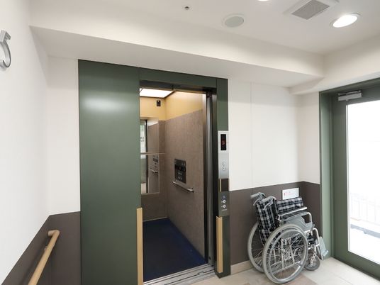 エレベーターが設置されている。緑色の枠の乗降口のすぐ横に、車椅子が折りたたまれた状態で置かれている。