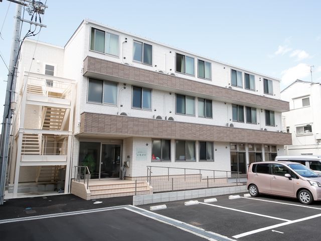 明るい日差しの建物外観 外観 清潔感のある白と茶色の外壁が印象的な建物です。ゆとりのある駐車スペースが設けられ、アクセスも良さそうです。