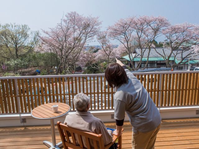 桜を見るテラスのシーン 入居者様 ケアを受ける方が、援助を受けながらテラスで穏やかなひとときを過ごしています。四季折々の景色をここから眺められる落ち着きのある空間です。