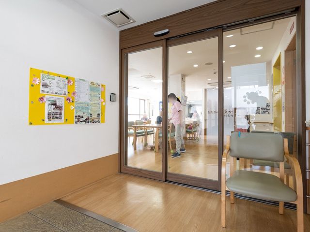 掲示板のある廊下 玄関 広々としたエントランスには掲示板が設置され、明るい食堂へと続くガラスの扉があります。扉は自動で開閉するため、すべての方が快適に移動できます。