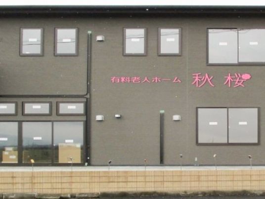 施設の建物の壁面には施設名がピンク色で表示されている。壁の色はグレー系で、多くの窓が設置されている。