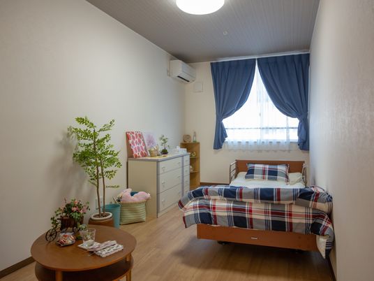 青いカーテンが掛けられている入居者様の居室。介護用ベッドと棚、ローテーブルと観葉植物が置かれている。