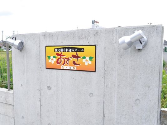 コンクリートの塀に、色鮮やかな施設の看板が掲げられている。看板の両脇には、ライトが一つずつ設置されている。
