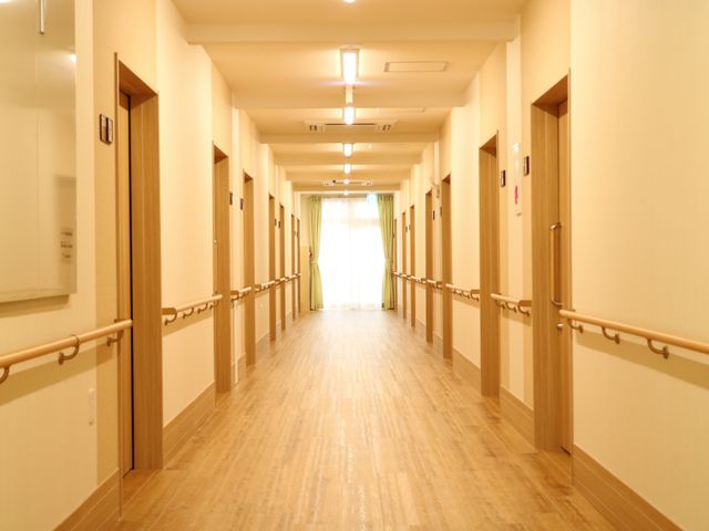 温かみのある廊下 廊下 床には木目調が施され、壁には手すりが設置されている、安全かつ心地よい移動スペースです。突き当りのカーテンからは自然光が差し込み、明るい雰囲気を演出しています。