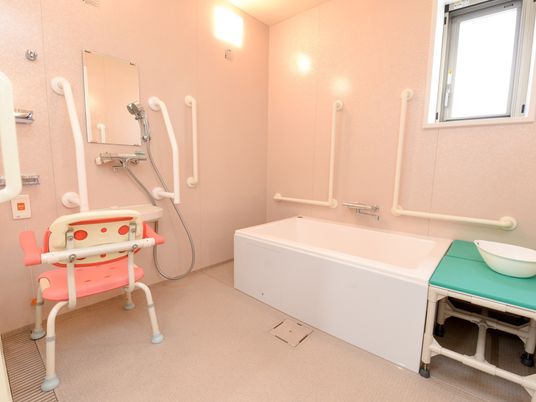 浴室のシャワースペースに、ピンク色のひじ掛けついた椅子が設置されている。壁には呼び出しボタンが取りつけられている。