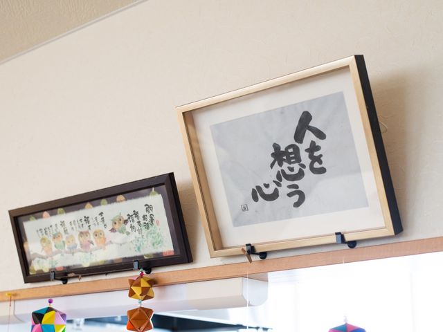 壁掛けの掲示物と書 展示物 和やかな文字が描かれた掲示物が、壁に飾られ心温まる雰囲気を醸し出しています。ここで働けば、日々の中に潤いを見つけられるかもしれません。