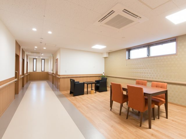 居心地の良い広々廊下 談話室 床には光沢があり、壁面はあたたかみのある色合いで、待合スペースも設けられています。清潔感があり、ゆったりとした通路は居心地が良さそうです。