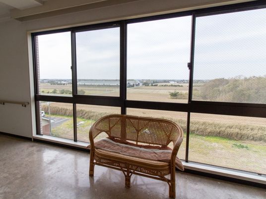 施設の写真 大きな窓のある廊下で、椅子が置かれている。建物周辺には高い建物がないので見晴らしが良く、周辺には田畑が広がっている。