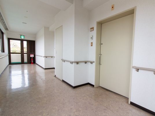 施設の写真 白を基調とした清潔感のある廊下で、居室の扉横にはインターホンが設置されている。非常扉の前には、消火器が置かれている。