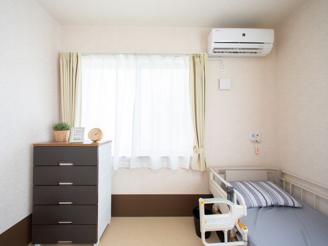 快適な居室の一角 居室 手元の時計が置かれたチェストと、優しい色合いのカーテンが心地よいプライベート空間です。安全手すり付きのベッドで、ゆったりとお過ごしいただけます。