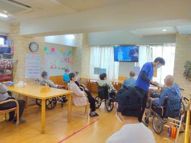 共有スペースでのひととき ラウンジ ゆったりとしたラウンジで、スタッフが入居者の方々と対話しています。テレビの前には様々な椅子が配され、視聴や交流が楽しめる空間です。