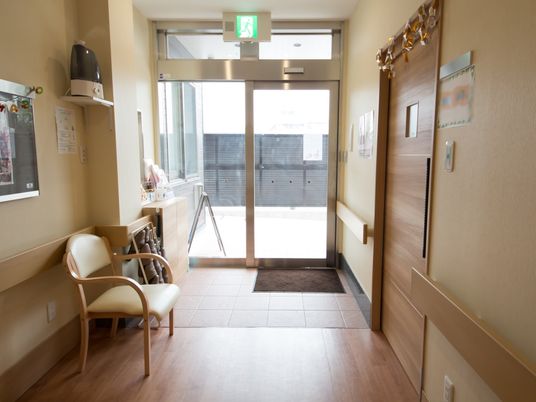 施設の写真 施設内の玄関は、段差のないフラットな床となっている。左側には、茶色いスリッパや椅子が1脚置かれている。