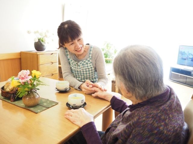 スタッフと利用者の交流スペース リビング 自然光が差し込むリビングで、スタッフが利用者と笑顔で交流しています。リラックスした雰囲気です。