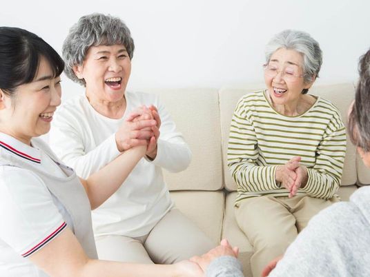 施設の写真 ベージュのソファを囲んで楽しそうに話している高齢女性たちと白衣を着た女性スタッフ。