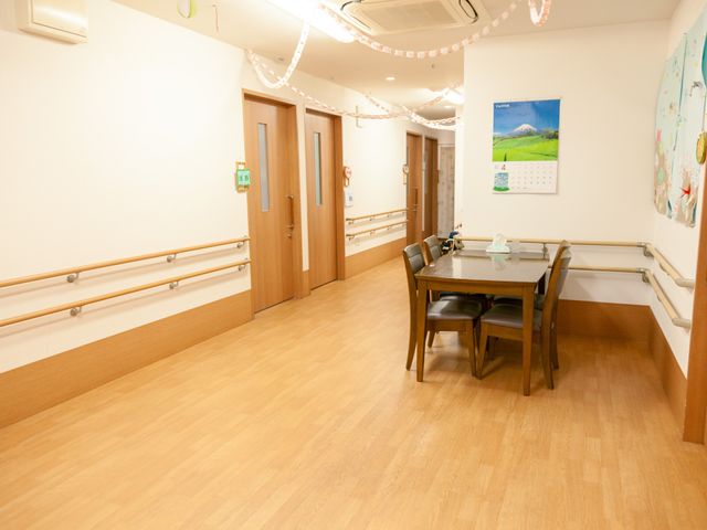 居心地の良い居室拝見 廊下 広々とした明るいスペースに、手すりが備えられた廊下が見えます。テーブルと椅子で構成されたくつろぎのコーナーでは、ゆっくりとお時間を過ごせそうです。
