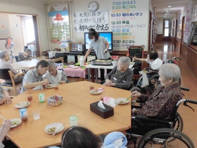 食事時の共有スペース リビング ゆとりある食堂で、スタッフが優しく配膳をしています。利用者の皆様は楽しそうに食事をされており、笑顔が見られます。