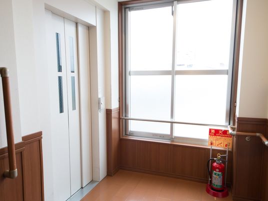 腰窓がある廊下の突き当りに設けられたエレベーターである。エレベーターホールには手すりが備わっている。