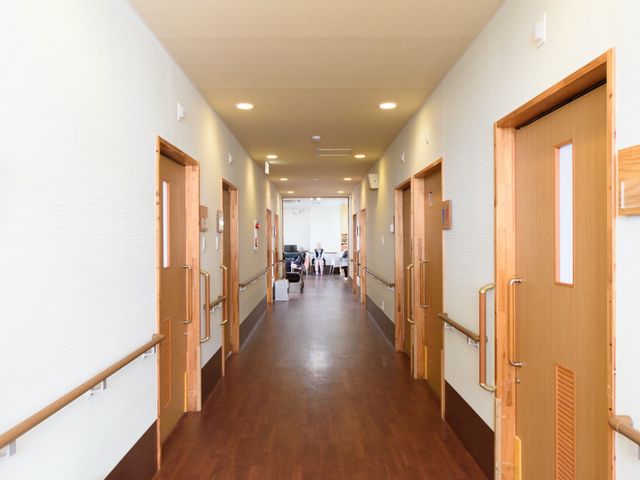 暖かみのある廊下 通路 広々とした廊下は手すりが設置され、明るい環境が整っています。廊下の先には居心地の良い共有スペースが見え、静かに過ごせる様子です。