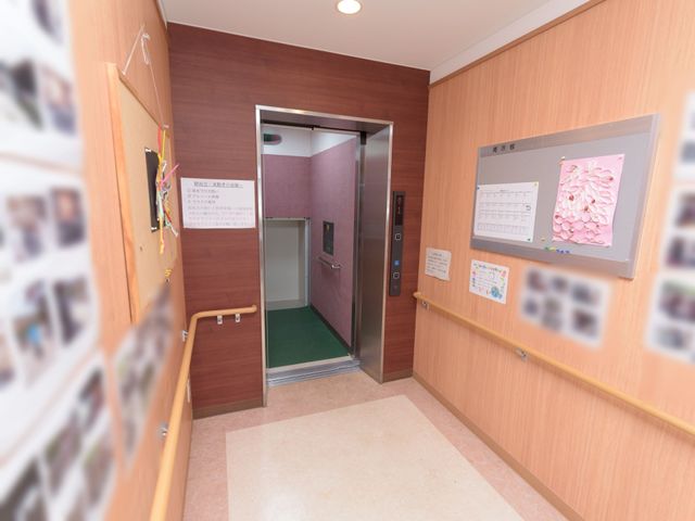 明るい廊下とエレベーター エレベーター 手すりが完備された廊下です。壁には掲示物や装飾があり、訪れる人々に安心感を提供しています。