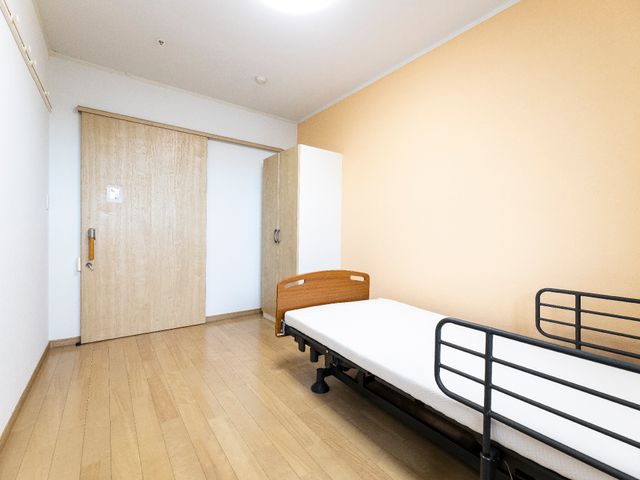 居室のベッドと扉 居室 シンプルなデザインの個室です。清潔感があり、日々の生活に必要な家具が整っており落ち着いた時間を過ごせる空間を提供しています。