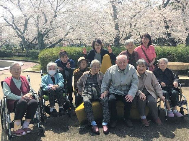 春の中庭で笑顔の集合 周辺環境 桜の下で談笑する利用者の方々、和やかな雰囲気の中庭でのひとときを垣間見ることができます。利用者の笑顔が輝く、温かな環境です。