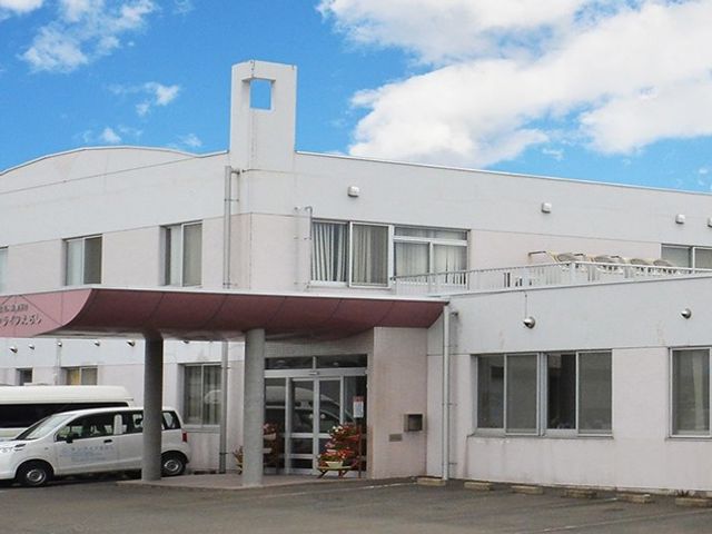 明るい建物外観 外観   青空の下にある明るい建物で、出入口前には車が停めやすいスペースがあります。入口まわりが見通しよく、来訪時の動線を確認しやすいです。