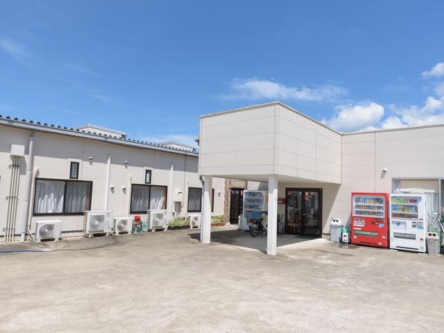 晴天下の白い建物外観 入口 快晴の下、清潔感のある白い建物が並ぶ広々としたエリアです。出入口に近く、自動販売機が設置されており、ひとときの休息にも適しています。