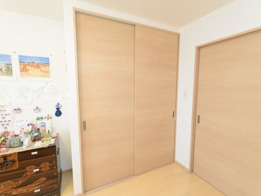フローリングの部屋に、膝丈くらいの和箪笥が置かれ、その上に人形や置物が飾られている。部屋のドアは木製の引き戸になっている。