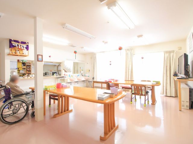 明るい共有スペース 食堂 広々とした食堂で、一人が車椅子でくつろぎながら過ごしています。テーブルや椅子が配置され、暖かな日差しの中で交流が生まれる場です。