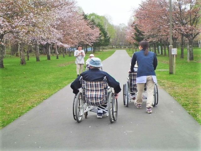 車椅子で散歩する人々 お散歩 緑豊かな中庭で、スタッフが車椅子を押して利用者様と散歩をしています。心地よい自然の中で、共に過ごす穏やかな時間があります。