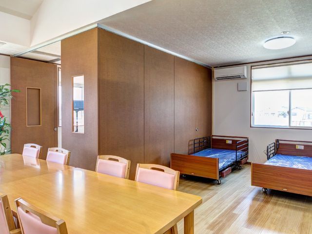 居室のベッドとテーブル 共有スペース 木製のベッドと大きなテーブルが配置された、明るく広々とした居室です。ゆったりとくつろげる空間が提供されています。