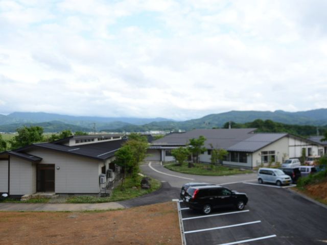 山並みを背景にした外観 外観 豊かな自然に囲まれ、穏やかな景色が広がる閑静なロケーションです。清潔感溢れる外壁と整備された駐車場が見受けられます。