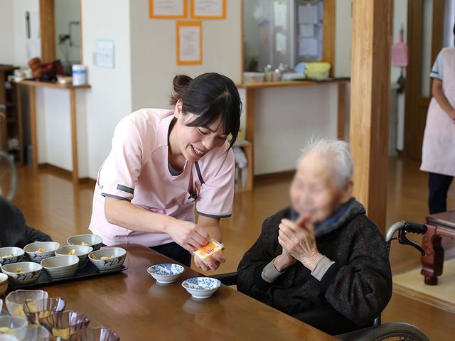 会話が弾むダイニング ダイニングルーム 職員が笑顔で入居者とコミュニケーションをとりながら、テーブルに食事を配っています。