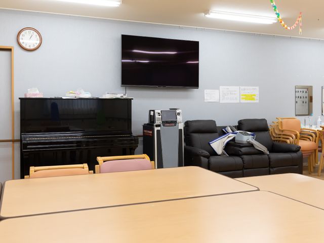 共用スペースとピアノ 共有スペース ゆったりとした共用スペースで、ピアノの音色に耳を傾けながらくつろぐことができます。壁掛けの大型テレビで、情報収集や娯楽をお楽しみいただけます。