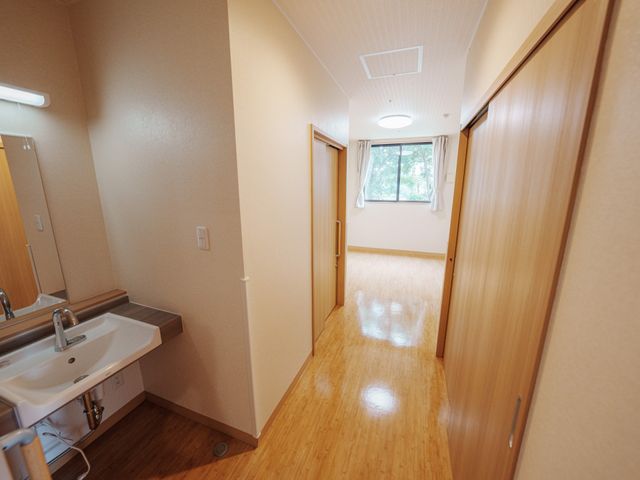 明るい個室のドアと洗面台 居室 ゆとりのある居室には、必要な備品が整い、窓からは自然光が差し込んでいます。快適な日々を過ごせる空間です。
