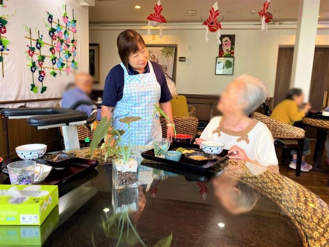 食堂でくつろぐ様子 食堂 明るい空間で、職員が利用者に温かく接しています。リラックスした雰囲気が感じられます。