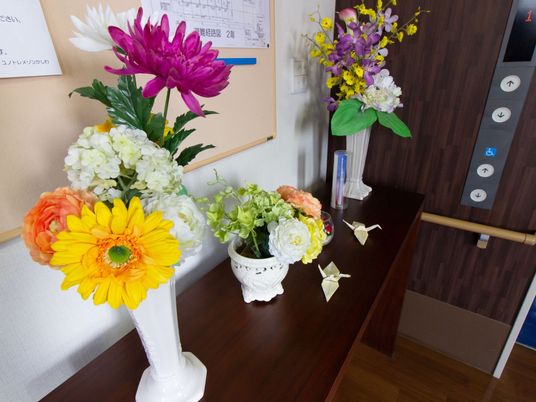 エレベーター乗降口の横に飾り棚が置かれている。その上に、色とりどりの花が生けられた花瓶が３つ載っている。