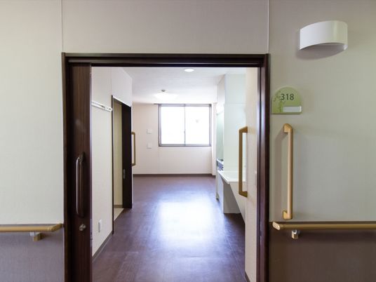 居室の入口から室内を見ると、正面に窓が見える。入口の両側すぐの所まで手すりが設置されている。緑色の板に居室番号が表示されている。