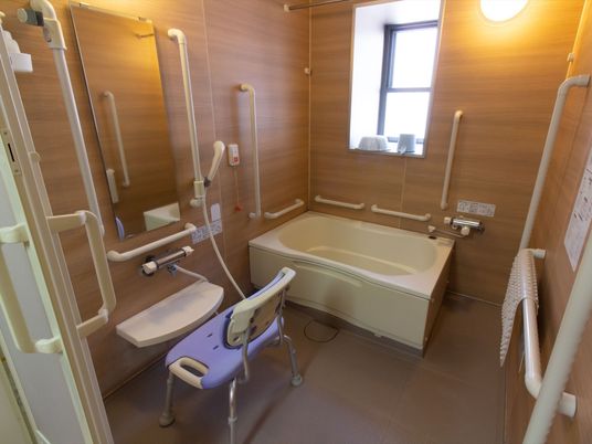 浴室は木目調の壁になっている。背もたれのある椅子が置かれている。滑り止めマットが手すりにかけて乾かされている。