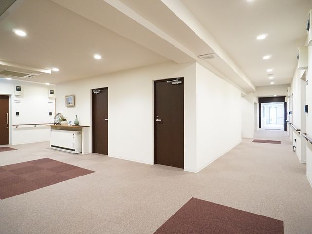明るく清潔な廊下 廊下 広々として明るい廊下は清掃が行き届いており、日々の業務に適した環境が整っています。左手には小物が飾られた棚が設置され、温かみのある雰囲気を演出しています。