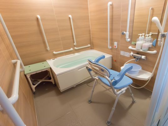 青いシャワー椅子が１台置かれた浴室。壁が木目調で温かみがあり、白い手すりが何本も備え付けられている。