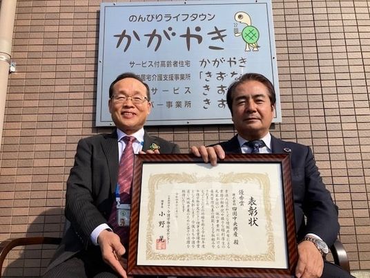 表彰状を持つ男性2人の看板前  