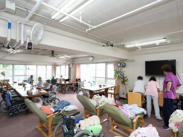 明るい広々とした食堂 ダイニング ゆとりある食堂では、明るい陽光が差し込み、職員が利用者の方々と会話を交わしながら過ごしています。多様な椅子やテーブルが配置され、テレビも備え付けられており、心地よい空間が広がっています。