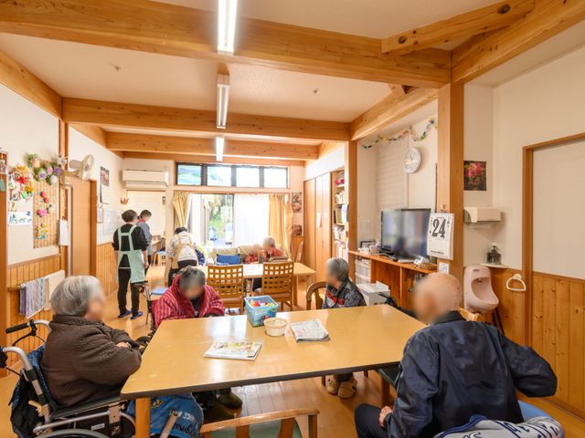 和やかな居室の様子 共有スペース 木の温もりあふれる居室で、利用者様が日常を優しく過ごしています。ケアスタッフが手厚くサポートしており、穏やかな雰囲気が感じられます。
