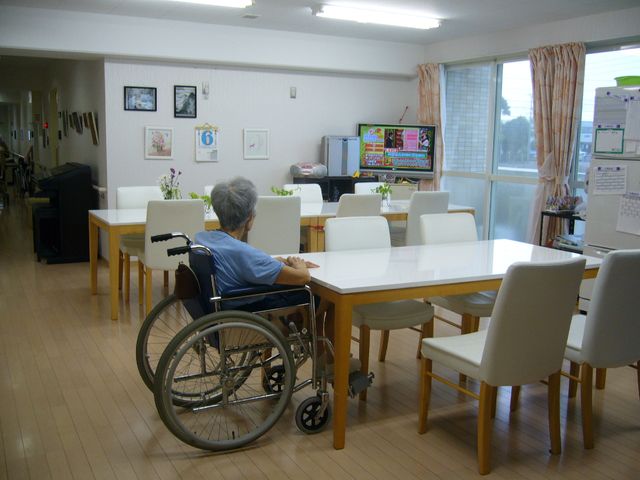 車椅子の人がいる食堂 共有スペース ゆったりとした食堂で、一人がくつろいでいる様子が伺えます。窓からの自然光が心地よい空間を演出しています。