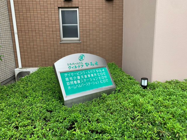 緑に囲まれた施設看板 外観   建物前に施設名の看板があり、植え込みに囲まれた落ち着いた外まわりです。出勤時の導線や周囲の雰囲気を確認しやすいです。