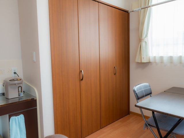 居室のクローゼットとテーブル 居室 落ち着いた色合いの居室には、収納豊富なクローゼットと広々とした作業スペースが備わっており、快適な生活空間を提供します。