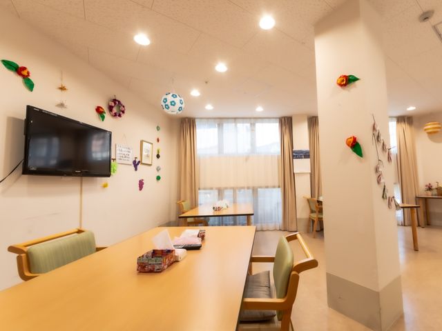 食堂のテーブルとテレビ 共有スペース 装飾が施された食堂で、テーブルには雑誌が置かれ、壁掛けのテレビも設置されています。ゆったりとした雰囲気の中で休憩が取れるスペースです。