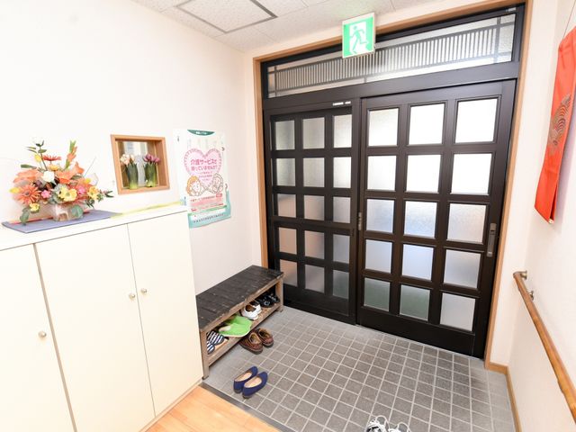 エントランスの角度から 玄関 おしゃれな花と掲示物が飾られた明るい入口部分です。手すりがあり、安全に配慮されている様子がうかがえます。